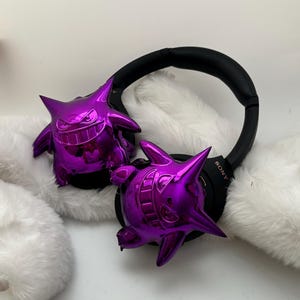 May include: Black Sony headphones with two shiny purple Gengar figures attached to the earcups. The Gengar figures have a glossy finish and are resting on a white, fluffy surface. The headphones are black.