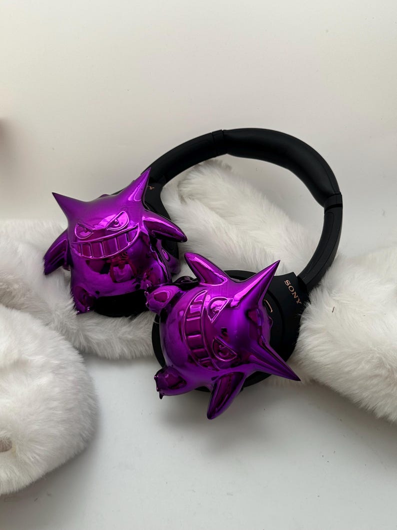 May include: Black Sony headphones with two shiny purple Gengar figures attached to the earcups. The Gengar figures have a glossy finish and are resting on a white, fluffy surface. The headphones are black.