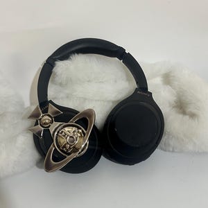 May include: Black Sony headphones with a silver and gold decorative accent. The headphones are resting on a white, fluffy surface. The accent features a planetary design.