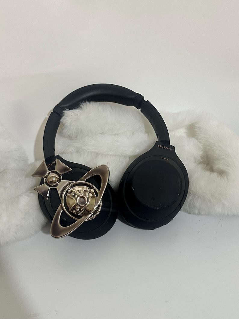 May include: Black Sony headphones with a silver and gold decorative accent. The headphones are resting on a white, fluffy surface. The accent features a planetary design.