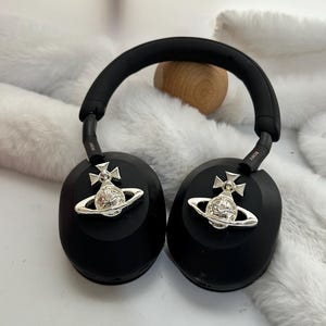 Puede incluir: Auriculares negros de diadema con adornos plateados de orbe y cruz. Los auriculares tienen una diadema y almohadillas acolchadas. La marca Sony es visible en los brazos. Los auriculares est&aacute;n sobre una superficie blanca y esponjosa.