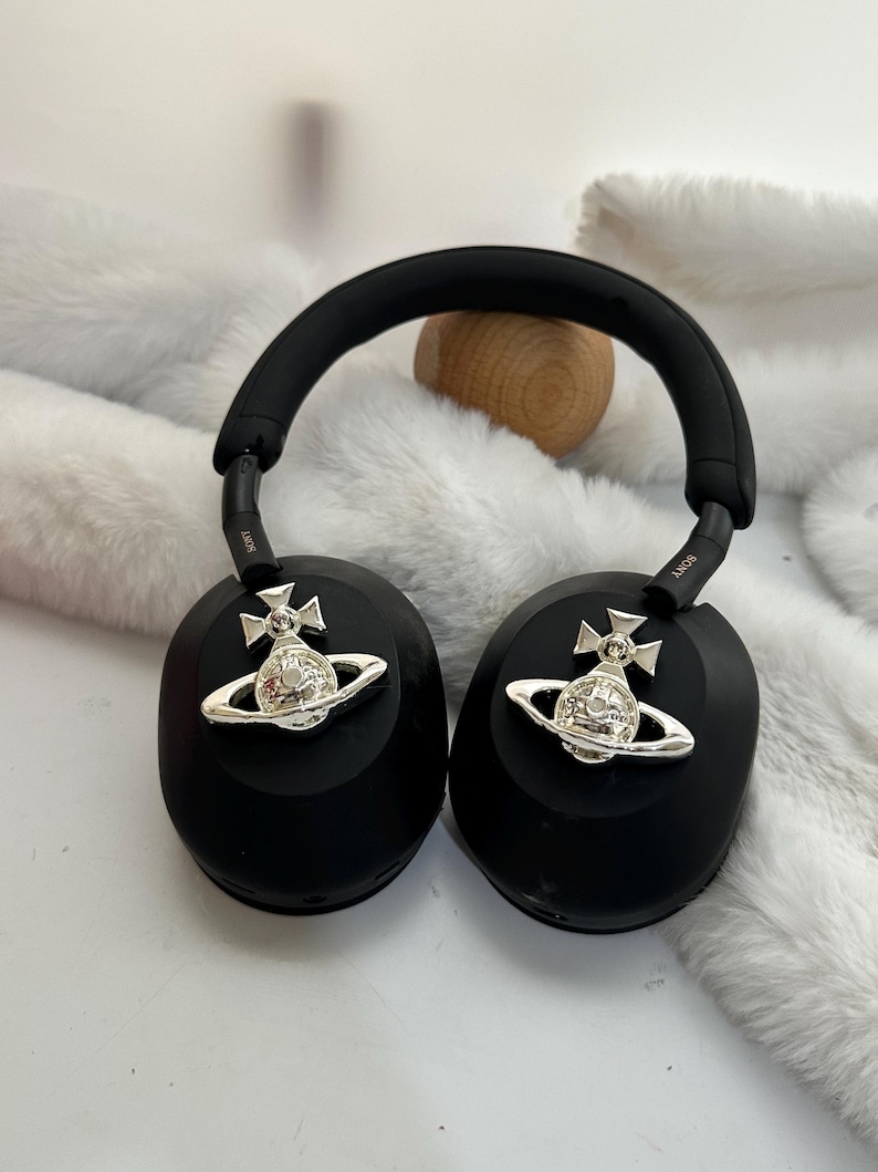 Puede incluir: Auriculares negros de diadema con adornos plateados de orbe y cruz. Los auriculares tienen una diadema y almohadillas acolchadas. La marca Sony es visible en los brazos. Los auriculares est&aacute;n sobre una superficie blanca y esponjosa.