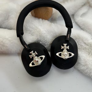Puede incluir: Auriculares negros de diadema con detalles plateados. Los auriculares tienen una diadema y almohadillas acolchadas. Cada auricular presenta un emblema plateado con un dise&ntilde;o de corona y orbe. La marca "SONY" es visible en los auriculares.