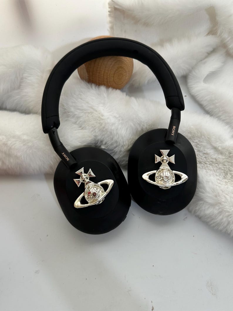 Puede incluir: Auriculares negros de diadema con detalles plateados. Los auriculares tienen una diadema y almohadillas acolchadas. Cada auricular presenta un emblema plateado con un dise&ntilde;o de corona y orbe. La marca "SONY" es visible en los auriculares.