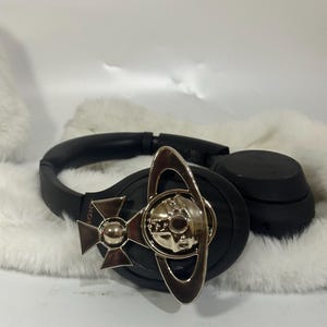 May include: Black over-ear headphones with a silver-toned decorative emblem. The emblem features a stylized orb design with a cross and ring elements. The headphones are resting on a white, fluffy surface.