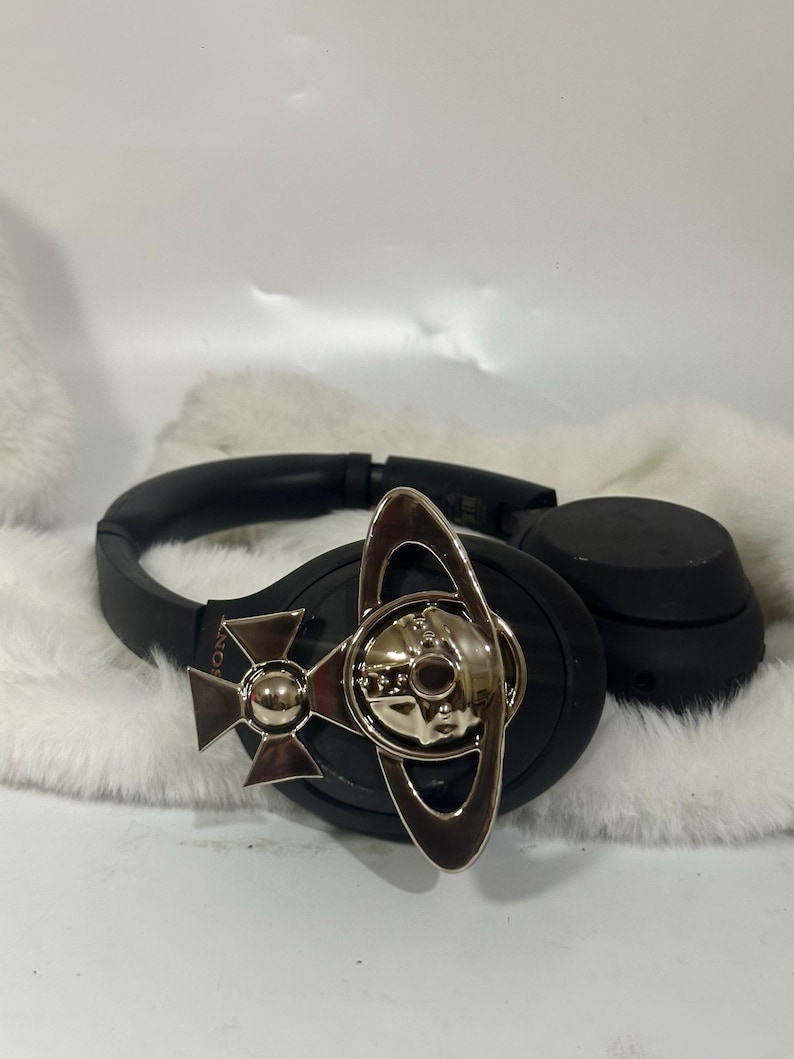 May include: Black over-ear headphones with a silver-toned decorative emblem. The emblem features a stylized orb design with a cross and ring elements. The headphones are resting on a white, fluffy surface.