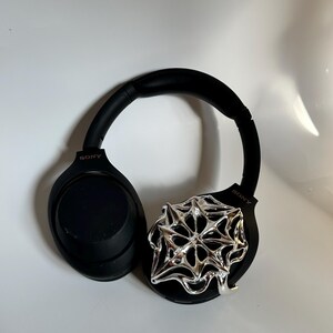 May include: Black Sony headphones with a silver, abstract, geometric design attached to the right ear cup. The headphones have a padded headband and earcups. The Sony logo is visible on both earcups.
