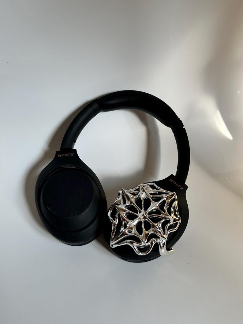 May include: Black Sony headphones with a silver, abstract, geometric design attached to the right ear cup. The headphones have a padded headband and earcups. The Sony logo is visible on both earcups.