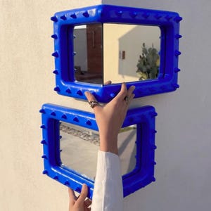 Puede incluir: Dos espejos de esquina rectangulares con un marco azul vibrante y una superficie texturizada con púas. Los espejos están diseñados para encajar perfectamente en la esquina de una pared, reflejando el entorno con una superficie de vidrio pulido.
