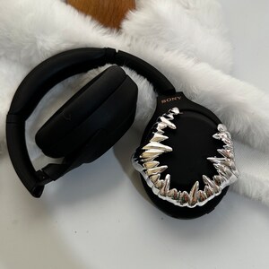 May include: Black wireless headphones with a silver, sharp, tooth-like design on the earcup. The text "SONY" is printed in pink on the earcup.