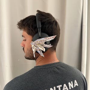 Puede incluir: Auriculares negros sobre la oreja con detalles decorativos plateados. La persona lleva una camiseta gris con la palabra "MONTANA" impresa en la espalda.