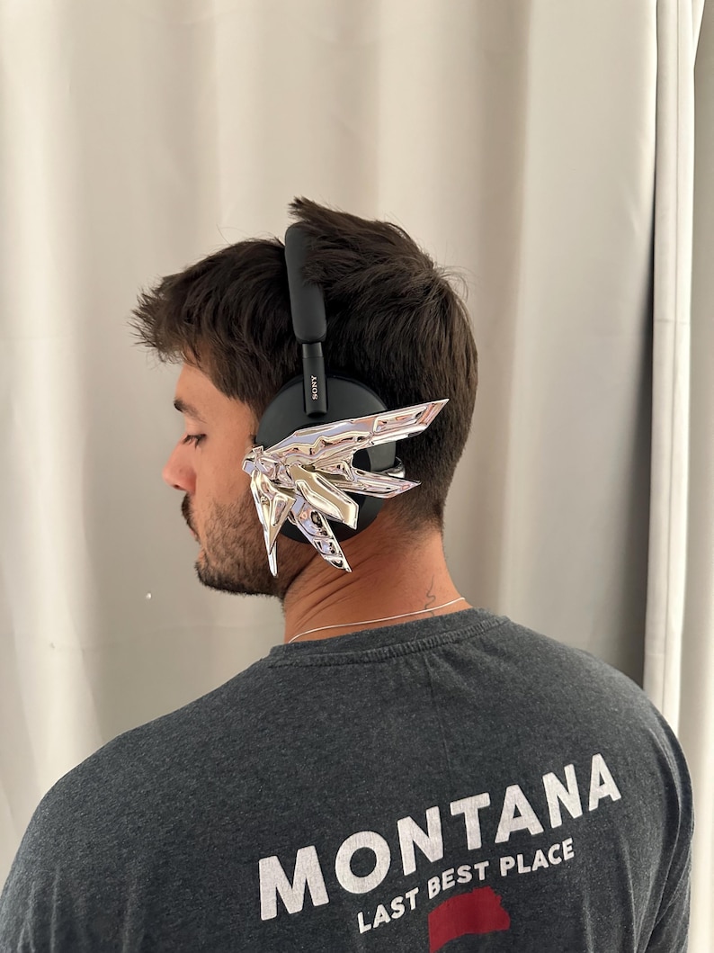 Puede incluir: Auriculares negros sobre la oreja con detalles decorativos plateados. La persona lleva una camiseta gris con la palabra "MONTANA" impresa en la espalda.