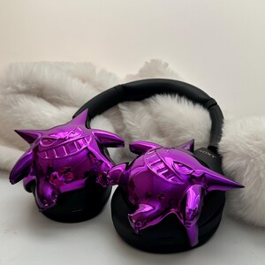 May include: Black headphones with shiny purple Gengar figures attached to the earcups. The headphones have a black headband and earcups. The Gengar figures are a vibrant purple color and have a glossy finish. The headphones are resting on a white, fluffy surface.