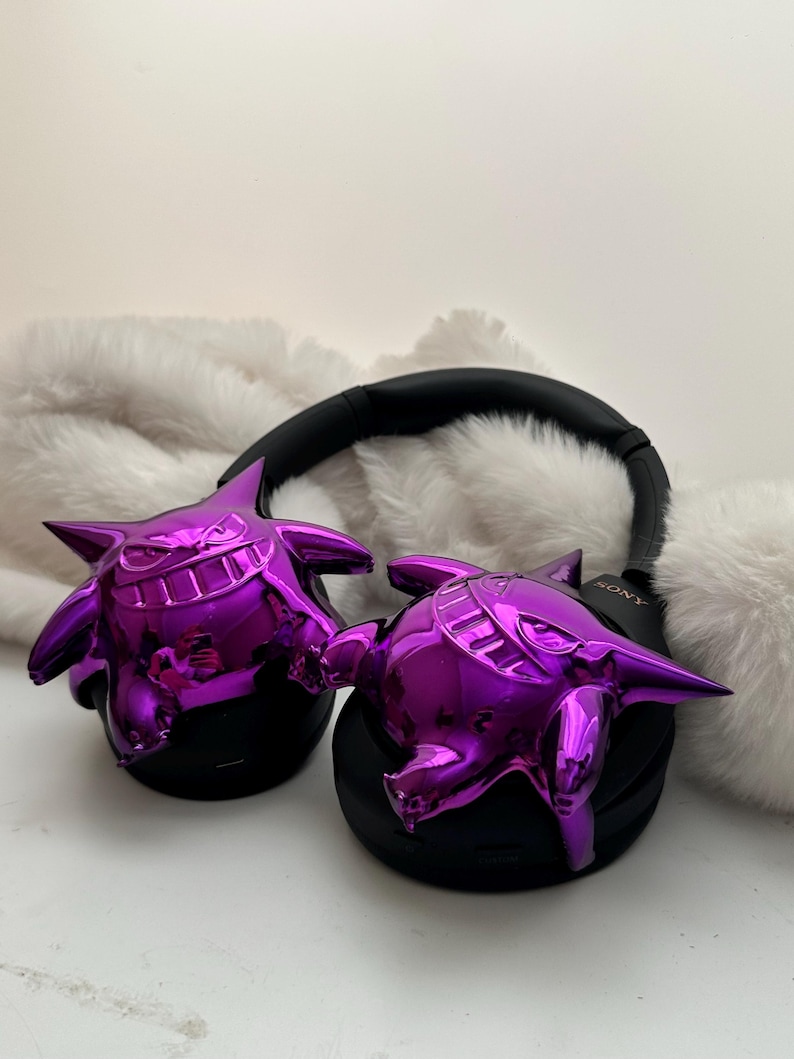 May include: Black headphones with shiny purple Gengar figures attached to the earcups. The headphones have a black headband and earcups. The Gengar figures are a vibrant purple color and have a glossy finish. The headphones are resting on a white, fluffy surface.