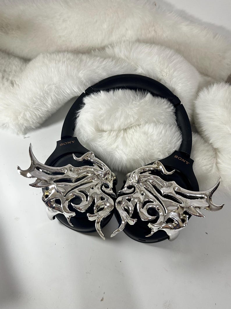 May include: Black Sony headphones with silver, ornate wing-shaped embellishments. The headphones rest on a white, fluffy surface. The earcups feature the Sony logo.