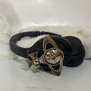 May include: Black over-ear headphones with a silver-colored decorative emblem. The headphones have a padded headband and earcups. The emblem features a stylized design with a skull and crossbones motif.