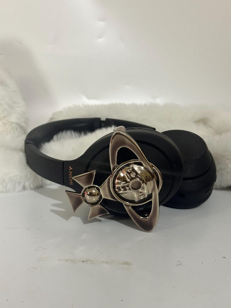 May include: Black over-ear headphones with a silver-colored decorative emblem. The headphones have a padded headband and earcups. The emblem features a stylized design with a skull and crossbones motif.
