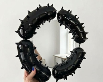 May include: A circular mirror framed by four glossy black, inflated, tubular segments. Each segment features a series of sharp, pointed spikes protruding from the surface, creating a textured, bold, and industrial appearance around the reflective glass.
