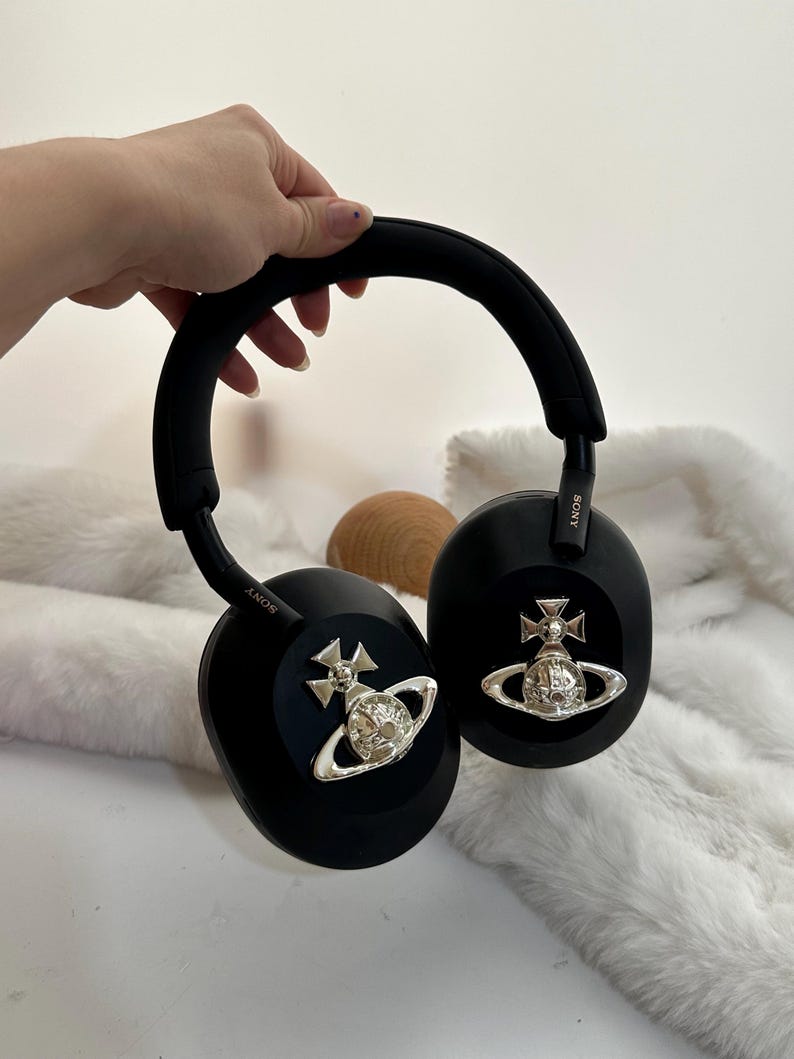 Puede incluir: Auriculares negros de diadema con detalles plateados. Los auriculares tienen una diadema y almohadillas acolchadas. Los auriculares presentan un dise&ntilde;o plateado de orbe y cruz. La marca "SONY" es visible en el lateral de los auriculares.