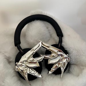 Puede incluir: Auriculares negros de diadema con adornos plateados abstractos en forma de mano en los auriculares. Los auriculares tienen una diadema y almohadillas negras. El fondo es una superficie blanca y esponjosa.