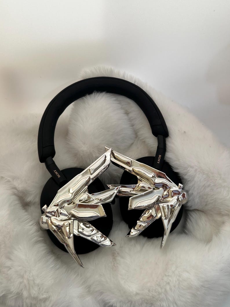 Puede incluir: Auriculares negros de diadema con adornos plateados abstractos en forma de mano en los auriculares. Los auriculares tienen una diadema y almohadillas negras. El fondo es una superficie blanca y esponjosa.