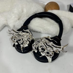 May include: Black over-ear headphones with silver decorative accents. The headphones have a black headband and earcups, with the brand name "SONY" visible. The silver accents feature an abstract, flame-like design. The headphones are on a white surface.