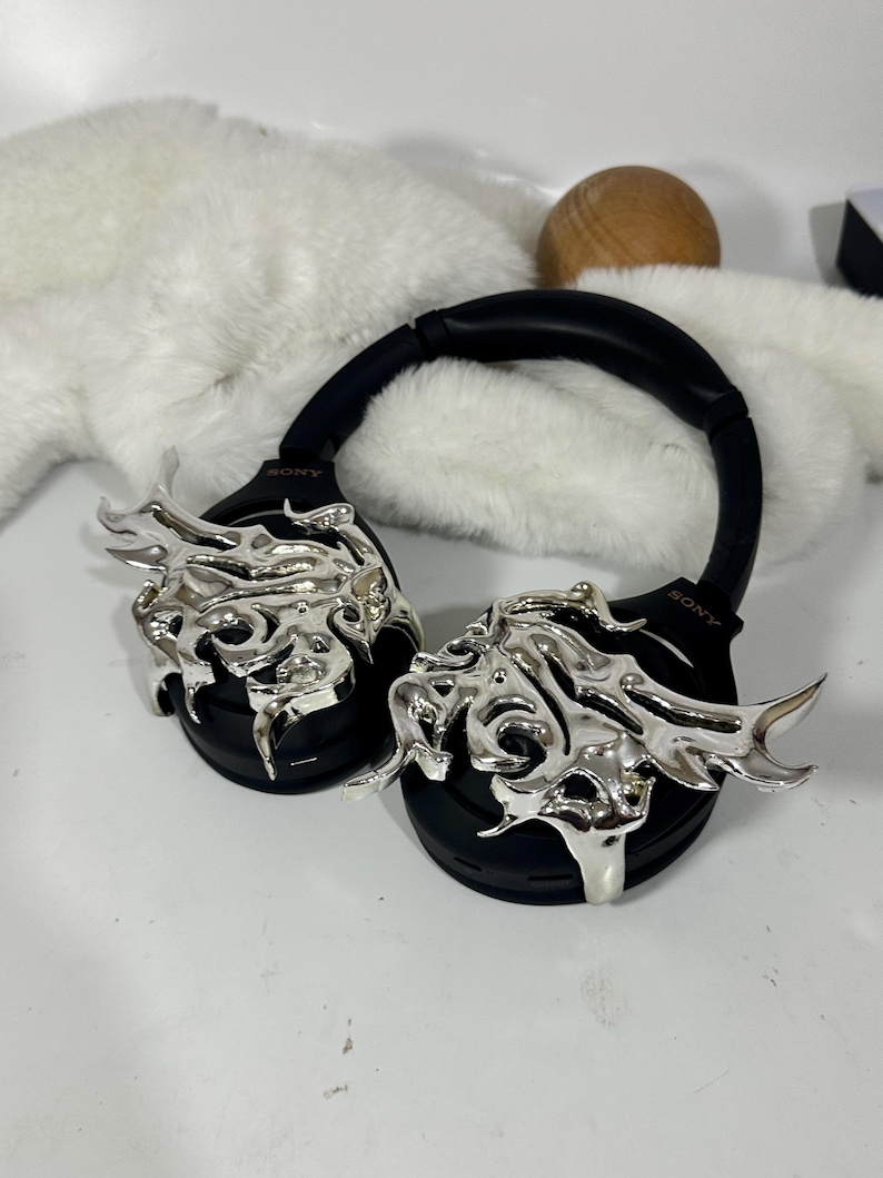 May include: Black over-ear headphones with silver decorative accents. The headphones have a black headband and earcups, with the brand name "SONY" visible. The silver accents feature an abstract, flame-like design. The headphones are on a white surface.
