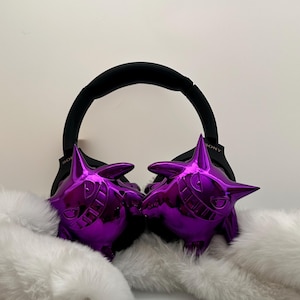 May include: Purple, glossy Gengar-themed earmuffs with a black headband. The earmuffs are set against a backdrop of fluffy white faux fur. The headband has the brand name "SONY" printed on it.