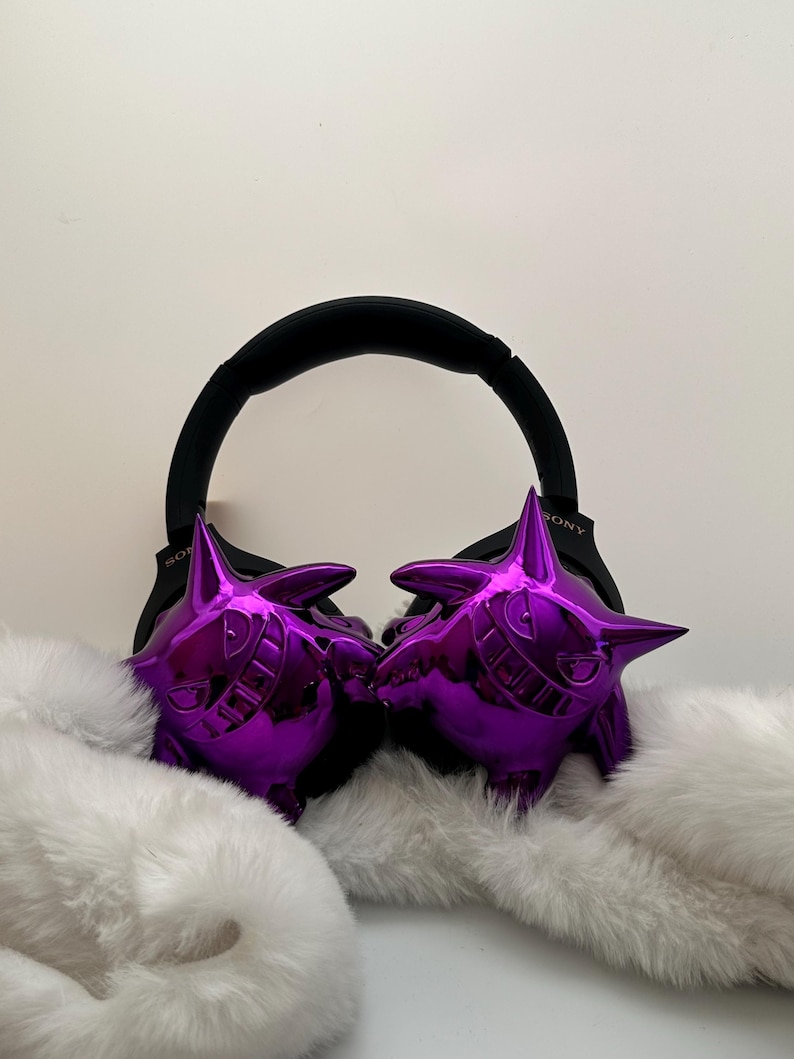 May include: Purple, glossy Gengar-themed earmuffs with a black headband. The earmuffs are set against a backdrop of fluffy white faux fur. The headband has the brand name "SONY" printed on it.