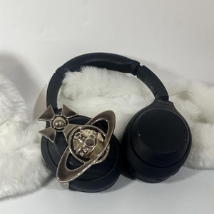 May include: Black over-ear headphones with a silver-colored decorative emblem. The emblem features a cross and a ring design. The headphones are resting on a white, fluffy surface.