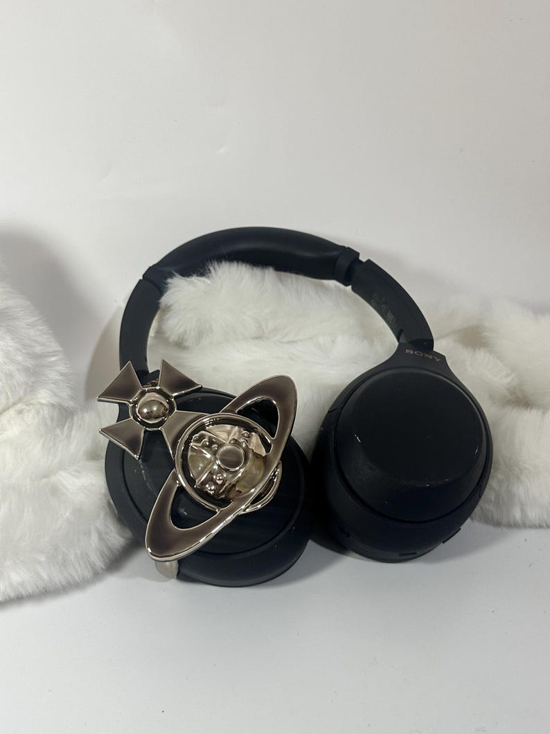 May include: Black over-ear headphones with a silver-colored decorative emblem. The emblem features a cross and a ring design. The headphones are resting on a white, fluffy surface.