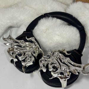 May include: Black over-ear headphones with silver flame-like embellishments. The headphones have the brand name "SONY" printed on the side. The background is a white surface with a fluffy white texture.