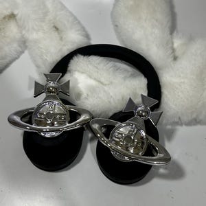 May include: A pair of black headphones with white fluffy earmuffs. The headphones have a silver orb with a cross on top and a ring around it.