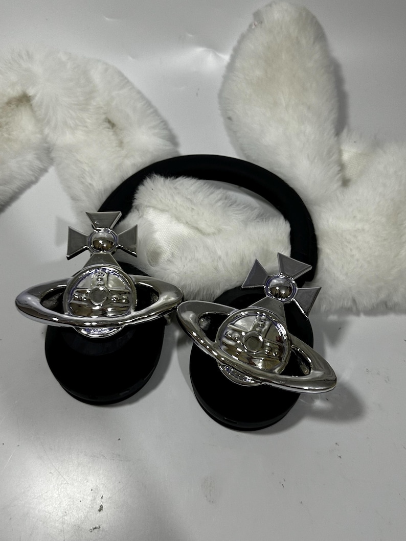 May include: A pair of black headphones with white fluffy earmuffs. The headphones have a silver orb with a cross on top and a ring around it.