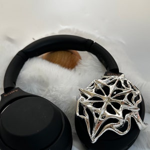 May include: Black Sony headphones with a silver spiderweb design on one ear cup. The headphones are resting on a white, fluffy surface. The Sony logo is visible on the headband.