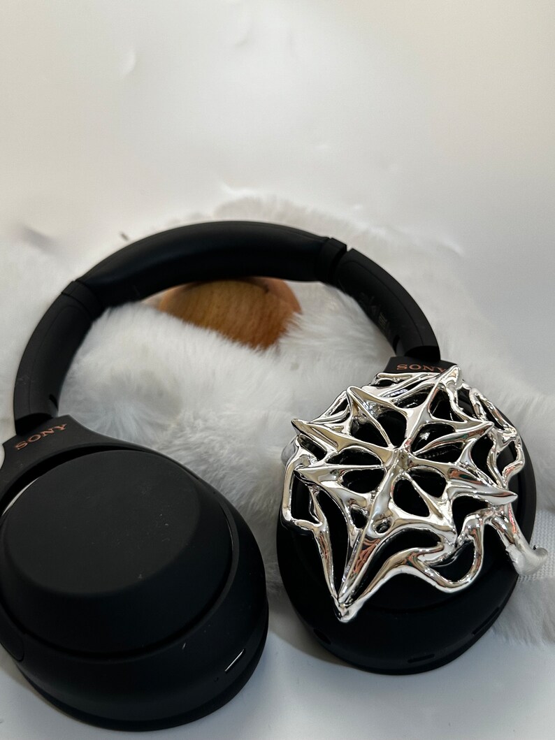 May include: Black Sony headphones with a silver spiderweb design on one ear cup. The headphones are resting on a white, fluffy surface. The Sony logo is visible on the headband.