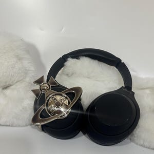 May include: Black Sony headphones with a silver-colored orb and cross pendant. The headphones are on a white surface with a fluffy white texture in the background. The pendant is a decorative accessory.