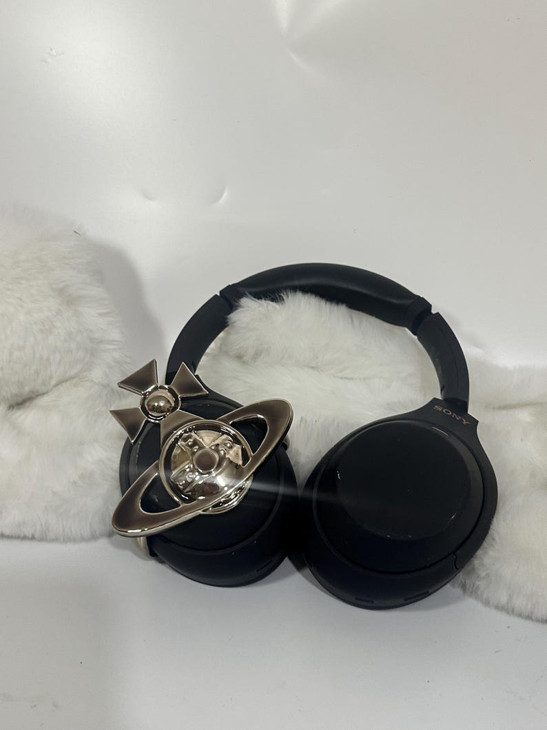May include: Black Sony headphones with a silver-colored orb and cross pendant. The headphones are on a white surface with a fluffy white texture in the background. The pendant is a decorative accessory.