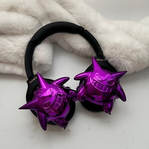 May include: Black Sony headphones with metallic purple Gengar figures attached to the earcups. The headphones rest on a white, fluffy surface. The Gengar figures have a shiny, reflective finish.