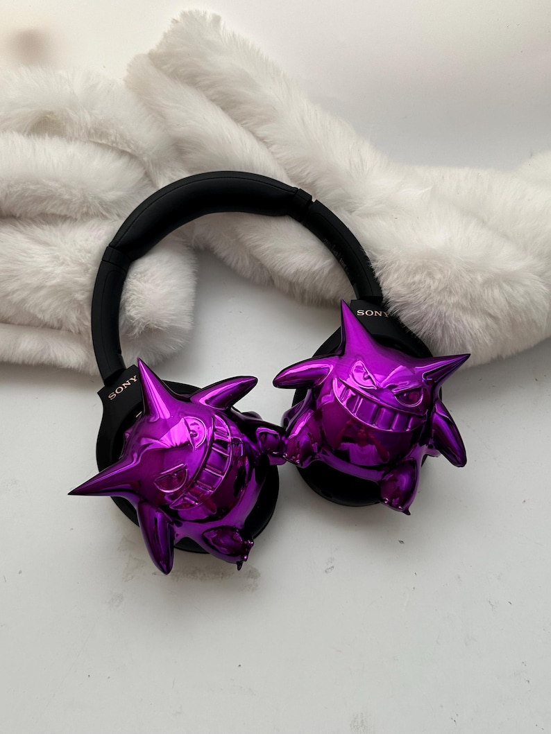 May include: Black Sony headphones with metallic purple Gengar figures attached to the earcups. The headphones rest on a white, fluffy surface. The Gengar figures have a shiny, reflective finish.