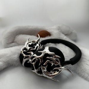 May include: Black headphones with silver, intricate, flame-like designs on the earcups. The headphones are on a white fuzzy surface.