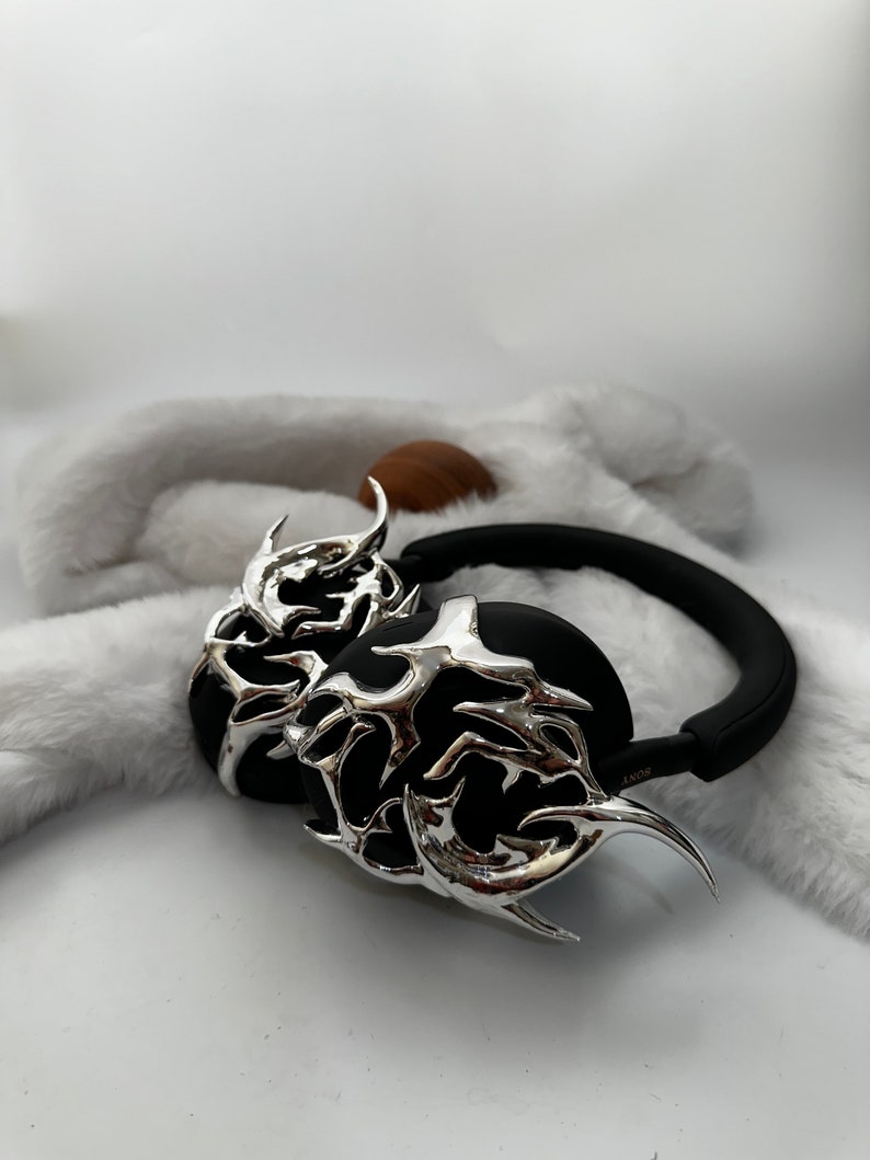 May include: Black headphones with silver, intricate, flame-like designs on the earcups. The headphones are on a white fuzzy surface.