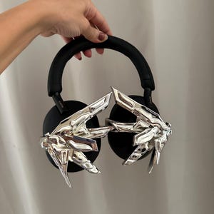 Puede incluir: Auriculares negros de diadema con detalles plateados en forma de manos. Los auriculares tienen una diadema y almohadillas acolchadas. Los detalles plateados son detallados y a&ntilde;aden un elemento de dise&ntilde;o &uacute;nico.