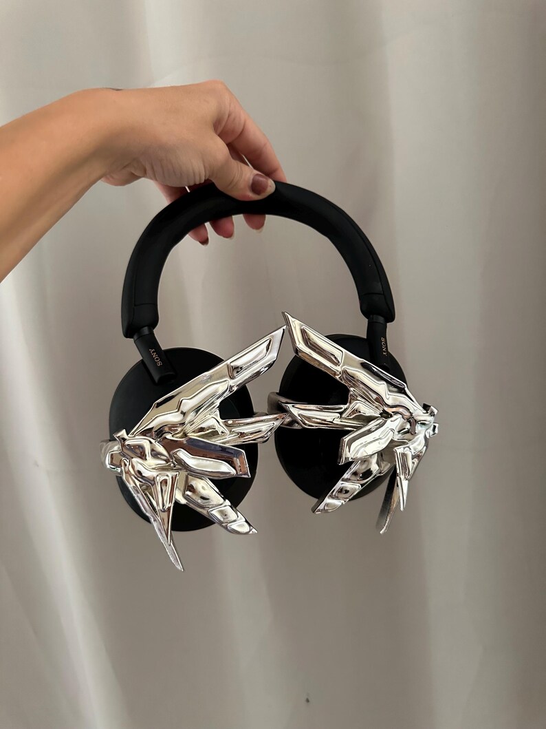 Puede incluir: Auriculares negros de diadema con detalles plateados en forma de manos. Los auriculares tienen una diadema y almohadillas acolchadas. Los detalles plateados son detallados y a&ntilde;aden un elemento de dise&ntilde;o &uacute;nico.