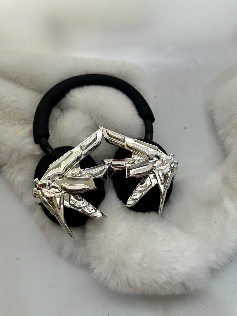 Puede incluir: Auriculares negros con detalles geom&eacute;tricos plateados. Los auriculares descansan sobre una superficie blanca y esponjosa. Las almohadillas y la diadema son negras.