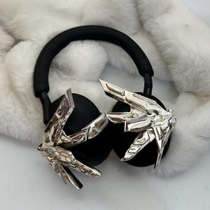 Puede incluir: Auriculares negros de diadema con detalles decorativos plateados. Los auriculares tienen una diadema y almohadillas negras. Los detalles plateados tienen un dise&ntilde;o angular y geom&eacute;trico. Los auriculares est&aacute;n sobre una superficie blanca y esponjosa.