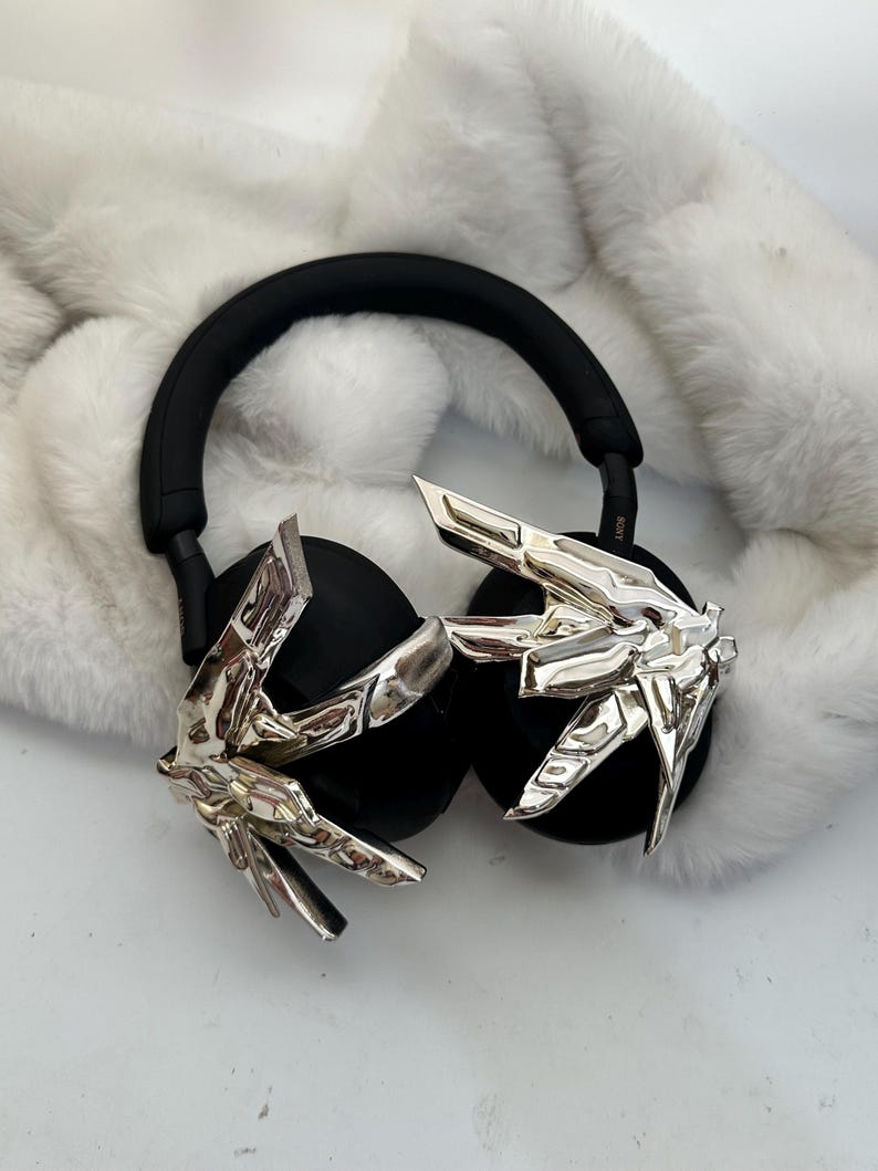 Puede incluir: Auriculares negros de diadema con detalles decorativos plateados. Los auriculares tienen una diadema y almohadillas negras. Los detalles plateados tienen un dise&ntilde;o angular y geom&eacute;trico. Los auriculares est&aacute;n sobre una superficie blanca y esponjosa.