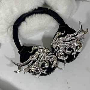 May include: Black Sony headphones with silver decorative accents. The headphones have a black headband and earcups. The silver accents feature an ornate, flowing design. The headphones are set against a white, fluffy background.