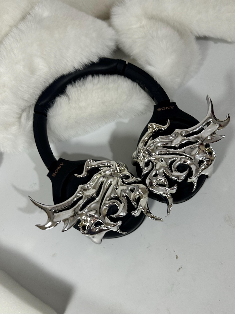 May include: Black Sony headphones with silver decorative accents. The headphones have a black headband and earcups. The silver accents feature an ornate, flowing design. The headphones are set against a white, fluffy background.