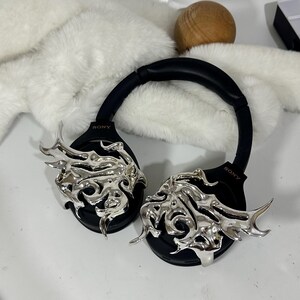May include: Black Sony headphones with silver decorative accents. The headphones have a black headband and earcups. The silver accents are an ornate, flame-like design. The headphones are on a white surface.